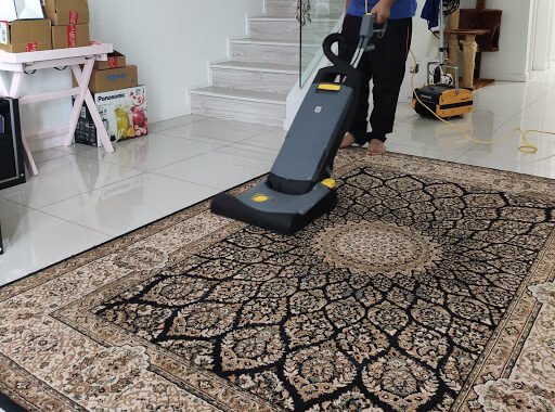 A patterned area rug being deep-cleaned with a commercial carpet vacuum cleaner in a bright, tiled living space with visible stairway and household items in the background